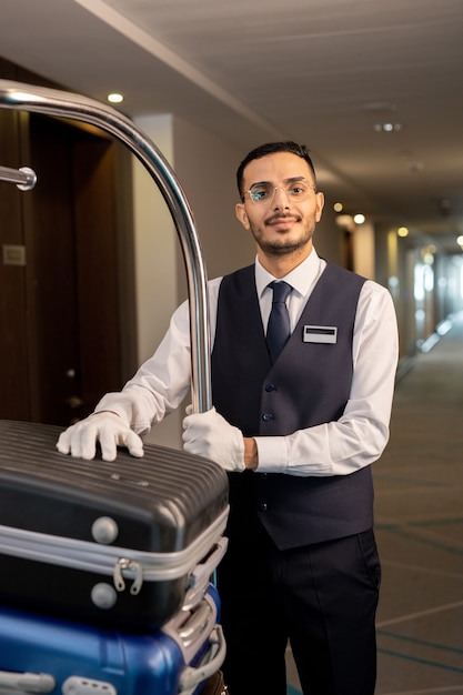 Junger ernsthafter Träger in Uniform und Brille, der mit Koffern von Gästen vor der Kamera im Korridor des zeitgenössischen Hotels steht