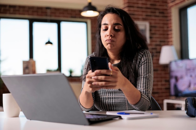 Junger Erwachsener, der auf dem Smartphone im Internet surft und die Social-Media-App verwendet, um SMS-Nachrichten in der Arbeitspause zu Hause zu senden. Arbeiten an Remote-Jobs mit Laptop und Mobiltelefon, Suche nach Online-Informationen.