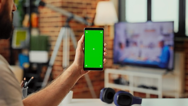 Junger Erwachsener hält Smartphone mit Greenscreen-Display im Wohnzimmer und verwendet isolierte Vorlage mit Chroma-Key und Attrappe. Analysieren des leeren Copyspace-Hintergrunds auf dem Handy.