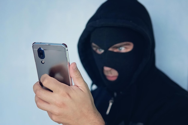 Junger Erwachsener in schwarzer Kleidung mit verstecktem Gesicht schaut auf den Smartphone-Bildschirm. Böswilliger Betrüger nutzt Mobilgeräte. Betrüger ruft an. Mobiler Schläger. Hacker entführt per Telefon. Handykontobetrug. Betrug.