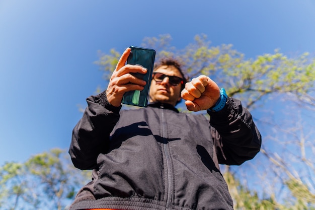 Junger erwachsener Mann, der das Mobiltelefon und die Smartwatch synchronisiert, um im Frühling oder Sommer bei Sonnenuntergang in einem Park Sport zu treiben. Konzept Sport Körperpflege und Technik