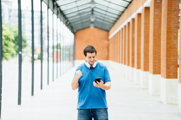 Junger erwachsener Mann, der das Smartphone überprüft und seine Leistung ausdrückt, die seine Faust glücklich auf der Straße ballt. Konzept gute Nachrichten erhalten Ziel erreichen