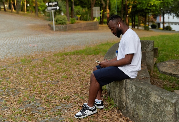 Junger erwachsener Mann, der sich nach einem Lauf mit einer Flasche Wasser und einem Smartphone-Outdoor-Training auf einem Park ausruht