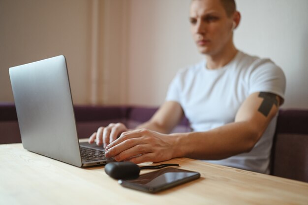 Junger europäischer Mann übergibt mit Smartphone und Kopfhörern, die an einem Laptop arbeiten