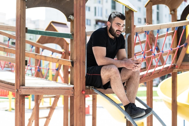 Junger fitter, gutaussehender erwachsener Mann, der sich im Freien auf dem Spielplatzpark ausruht