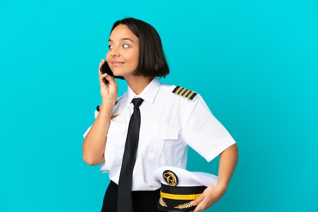 Junger Flugzeugpilot über isoliertem blauem Hintergrund, der ein Gespräch mit dem Mobiltelefon mit jemandem führt