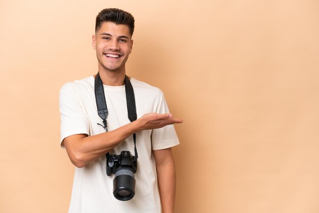 Junger Fotograf, kaukasischer Mann isoliert auf beigem Hintergrund, der eine Idee präsentiert, während er lächelnd in Richtung schaut