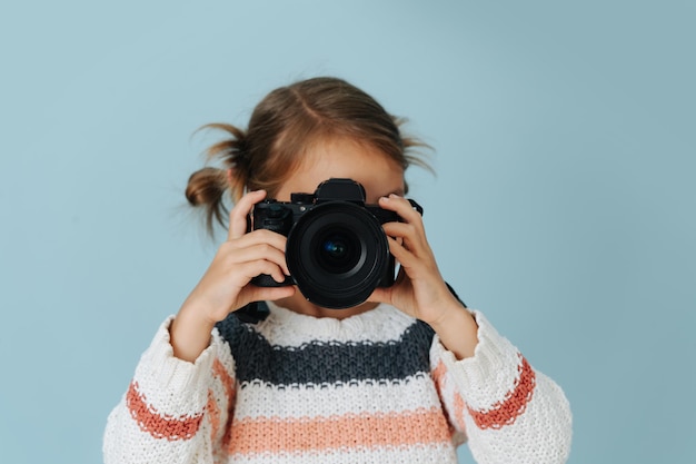 Junger Fotograf. kleines Mädchen in gestreiftem Pullover mit Haaren in Brötchen auf blauem Hintergrund. Sie fokussiert ihre Digitalkamera.