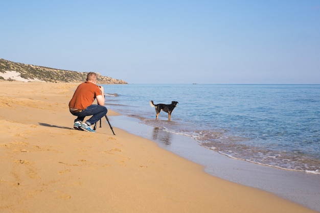 Junger Fotograf und Videofilmer, der mit der Kamera auf einem Stativ Fotos und Videos von Meer und seinem Hund macht. Mittelmeer.