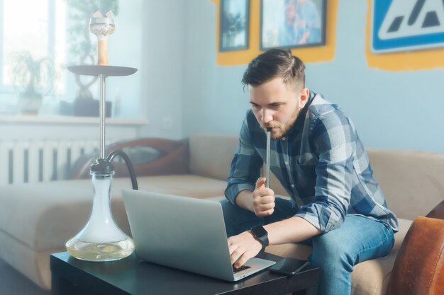 Junger Freiberufler, der eine Shisha raucht, während er von zu Hause aus arbeitet