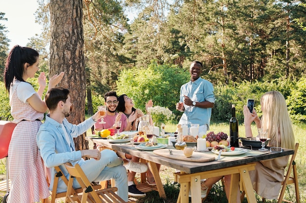 Junger freudiger Mann der afrikanischen ethnischen Zugehörigkeit, der durch festlichen Tisch vor seinen Freunden nach dem Abendessen im Freien tanzt, während eines der Mädchen Video aufzeichnet