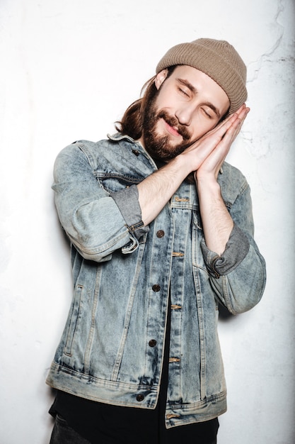 Junger fröhlicher bärtiger Hipster-Mann mit Hut in Jeansjacke, der auf Händen über der Wand schläft