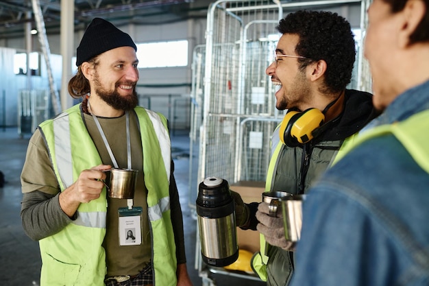 Junger, fröhlicher Ingenieur mit einer Tasse Thermoskanne, der lacht, während er mit einem Kollegen spricht