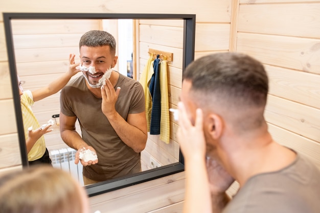 Junger fröhlicher Mann, der Schaum auf den Bart aufträgt, während seine süße kleine Tochter ihm bei der Morgenhygiene hilft, beide stehen vor dem Spiegel