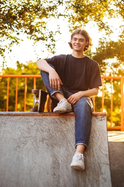 Foto junger, fröhlicher skater, der glücklich mit skateboard im modernen skatepark in die kamera schaut