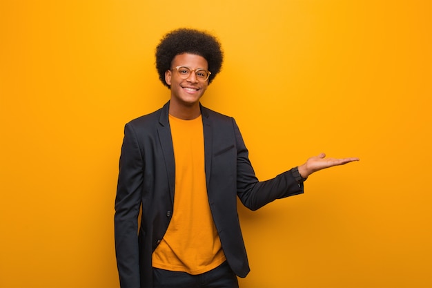 Junger Geschäftsafroamerikanermann über einer orange Wand, die etwas mit der Hand hält