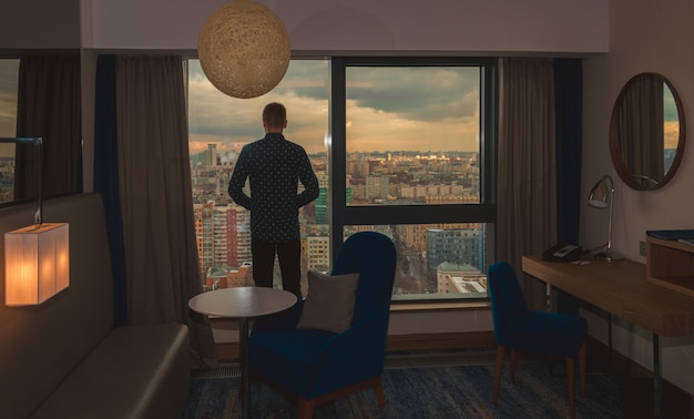 Junger Geschäftsmann auf dem Hintergrund der Panoramafenster mit dem Blick auf die Stadt vom Hotelzimmer zur Sonnenuntergangszeit