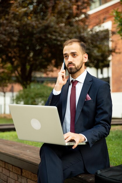 Junger Geschäftsmann, der am Laptop arbeitet und in einer Grünfläche telefoniert. Anzug und Business-Attitüde