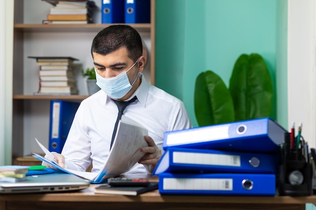 Junger Geschäftsmann, der am Tisch sitzt und Dokumente im Büro mit medizinischer Schutzmaske und Handschuhen liest