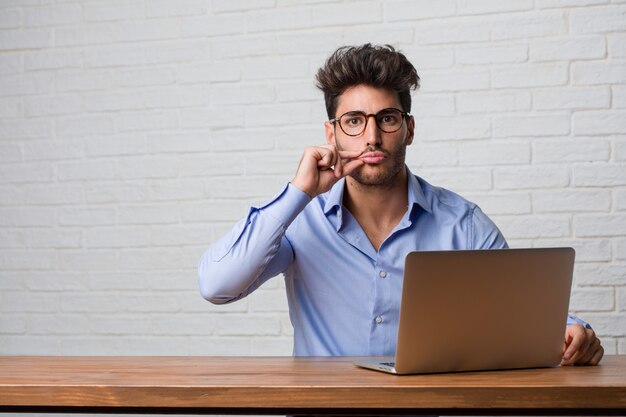 Junger Geschäftsmann, der an einem Laptop sitzt und arbeitet und ein Geheimnis hält oder um Ruhe bittet