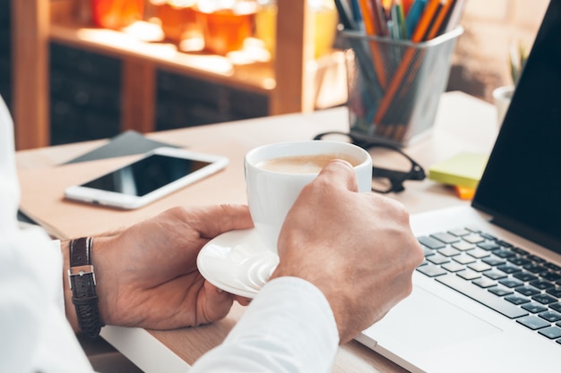 Junger Geschäftsmann, der an Laptop-Computer mit heißem Kaffee in der Hand arbeitet.