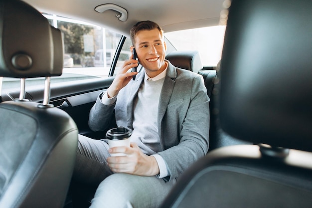 Junger geschäftsmann, der auf einem handy spricht und kaffee trinkt, während er auf dem rücksitz eines autos sitzt