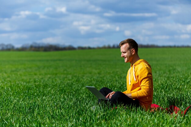 Junger Geschäftsmann, der auf grünem Gras sitzt und Laptop-Computer verwendet. Gut aussehender Mann, der am sonnigen Sommertag mit Computer im Park arbeitet. Naturreise und Entspannung im Freien. Freiberufliches Arbeitskonzept