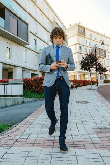 Junger Geschäftsmann, der die Straße hinuntergeht, während er auf das Handy schaut