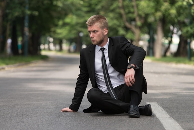 Junger Geschäftsmann, der draußen auf Asphalt im Park sitzt, während er sich von der Büroarbeit ausruht