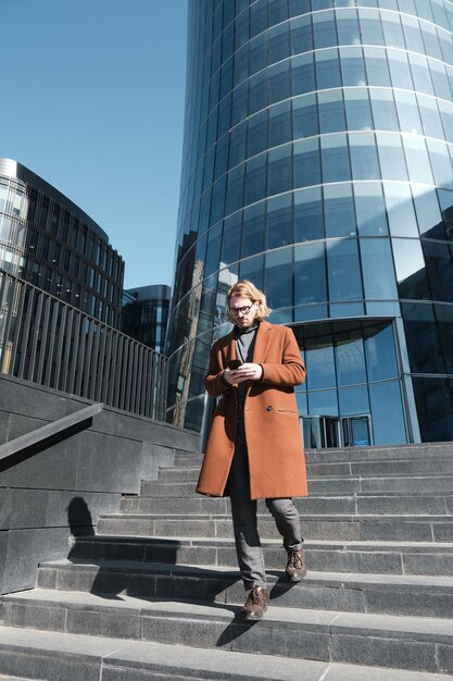 Junger Geschäftsmann, der Handy benutzt, während er die Treppe mit modernem Gebäude im Hintergrund hinuntergeht