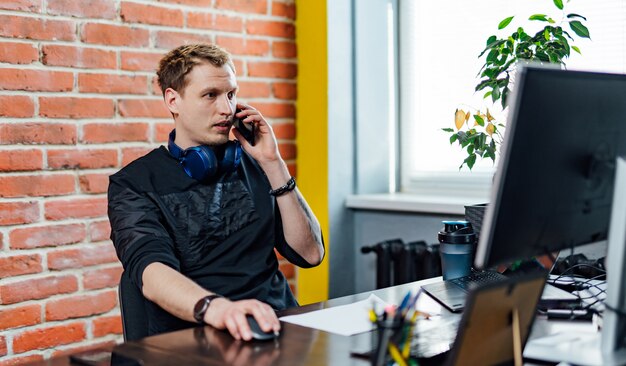 Junger Geschäftsmann, der im Büro arbeitet. Hübscher Geschäftsmann unter Verwendung des Computers und Unterhaltung am Handy beim Arbeiten im Büro