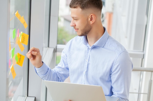 Junger Geschäftsmann, der mit einem Laptop in seinem Büro nahe dem Hintergrund eines Fensters steht