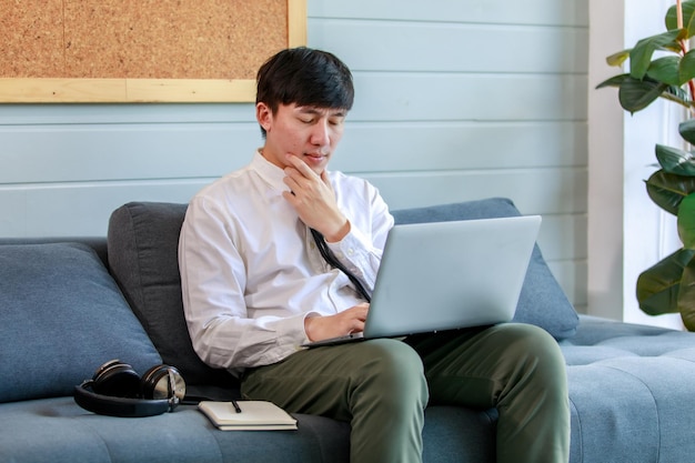 Junger Geschäftsmann, der mit Laptop-Kopfhörern und Papiernotizen auf der Ruhecouch der Bürolobby arbeitet und die Augen schließt, um eine Lösung für die Marketingstrategie und das Planungsnetzwerk zur Lösung schwieriger Aufgaben zu finden