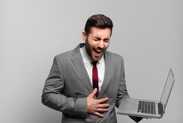 Junger Geschäftsmann, der über einen lustigen Witz laut lacht, sich glücklich und fröhlich fühlt, Spaß hat und einen Laptop hält