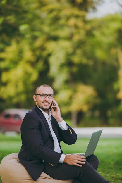 Junger Geschäftsmann im weißen Hemd, im klassischen Anzug, in der Brille. Mann sitzt auf weichem Hocker, telefoniert, arbeitet am Laptop-PC im Stadtpark auf grünem Rasen im Freien in der Natur. Mobiles Büro, Geschäftskonzept.