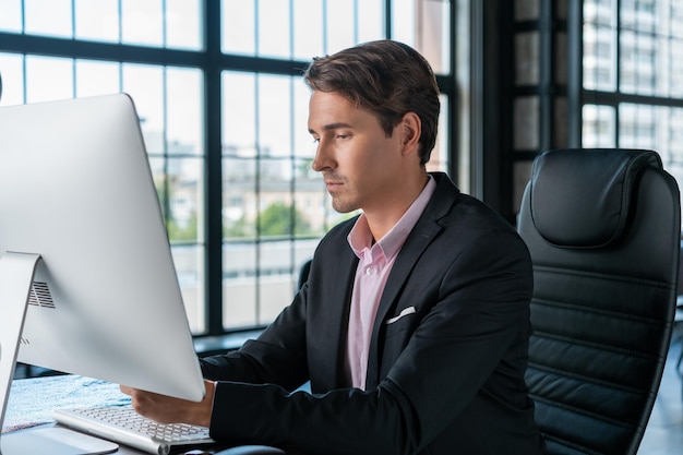 Junger Geschäftsmann in schwarzem Anzug und rosafarbenem Hemd, der am Tisch mit Computer sitzt Büroleiter, der in einem modernen Bürogebäude mit Glasfenstern arbeitet, verschwommenes Hintergrundkonzept der Arbeit