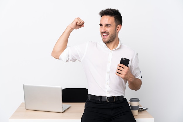 Junger Geschäftsmann mit einem Handy an einem Arbeitsplatz, der einen Sieg feiert