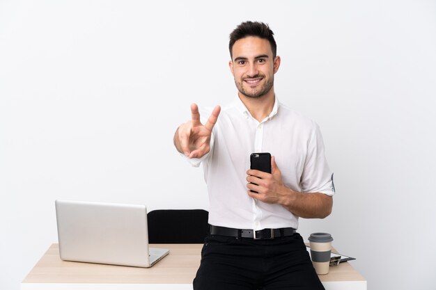 Junger Geschäftsmann mit einem Handy an einem Arbeitsplatz lächelnd und Siegeszeichen zeigend
