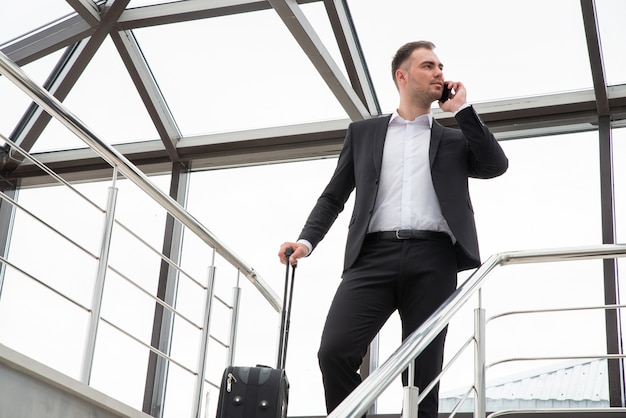 Junger Geschäftsmann mit Koffer im stilvollen Anzug, der durch Telefon spricht, während durch Treppen im modernen Geschäftsgebäude geht