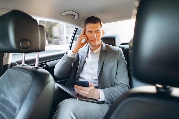 Junger Geschäftsmann müde von der Arbeit, die Hand an den Kopf hält, während er auf dem Rücksitz des Autos sitzt