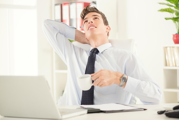 Junger Geschäftsmann sitzt im Büro, entspannt sich und trinkt einen Kaffee.