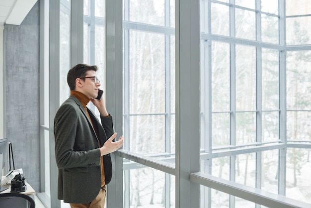 Junger Geschäftsmann steht vor dem Fenster und telefoniert während der Arbeit im Büro