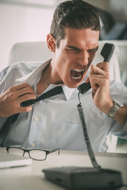 Junger gestresster Geschäftsmann, der im Büro sitzt und am Telefon schreit.