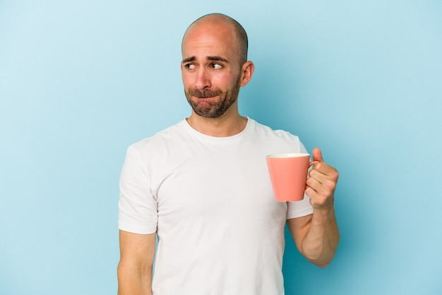 Junger glatzköpfiger Mann, der einen Becher auf blauem Hintergrund isoliert hält, verwirrt, fühlt sich zweifelhaft und unsicher.