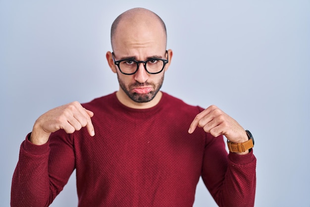 Junger glatzköpfiger Mann mit Bart, der über weißem Hintergrund steht und eine Brille trägt, die nach unten zeigt und traurig und verärgert aussieht, mit den Fingern die Richtung anzeigt, unglücklich und deprimiert.