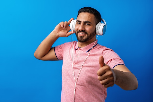 Junger glücklicher arabischer Mann mit Kopfhörern, die Musik vor blauem Hintergrund hören