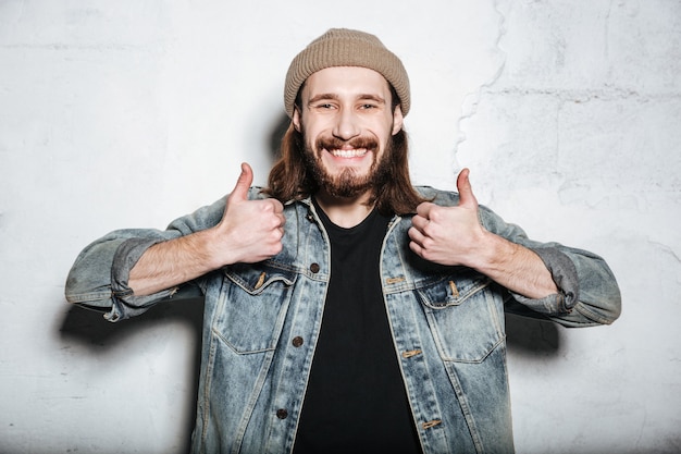 Junger glücklicher bärtiger Hipster-Mann mit Hut in Jeansjacke, der mit Daumen nach oben über der Wand posiert