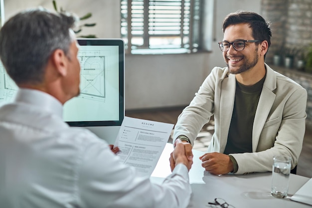 Junger glücklicher Kandidat und Mitglied des Personalteams beim Händeschütteln nach einem Vorstellungsgespräch im Büro