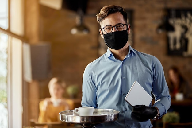 Junger glücklicher Kellner, der eine schützende Gesichtsmaske trägt, während er nach der Coronavirus-Epidemie in einem wiedereröffneten Café arbeitet