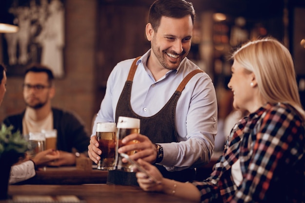 Junger glücklicher Kellner, der mit einer Frau spricht, während er ihr ein Glas Bier in einer Kneipe gibt